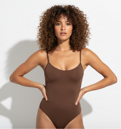 Professional 1:1 studiofoto van dezelfde vrouw. Geen verandering in gezicht of lichaam. Real skin texture. Real pores. Natural imperfections. Dunne schouderbandjes. Shapewear bodysuit in bruin. Clean white studio. Zachte schaduw. Direct eye contact. Pose handen in de zij met een lichte heupshift. Tan huid, curly hair shoulder length, vol, 28 jaar, sterke rustige blik.